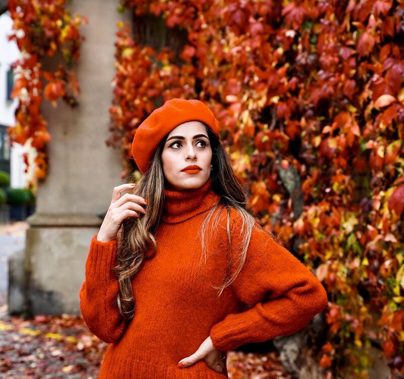 Roopsi Bansal in a rust-orange turtleneck sweater and matching beret, standing in front of a wall of red autumn ivy