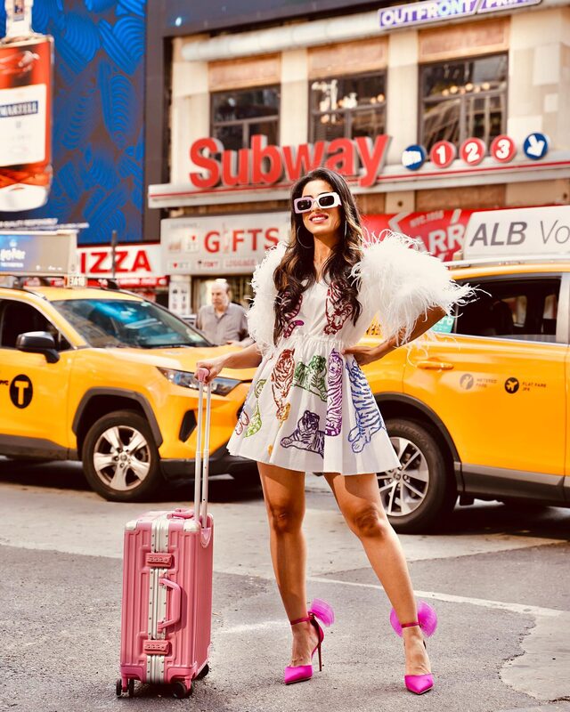 Roopsi Bansal in a white tiger-embroidered feather-sleeve dress with a rose pink AnyZip carry-on suitcase in Times Square, New York City