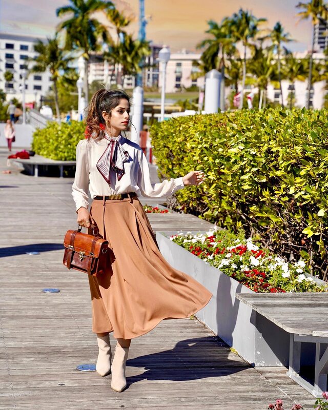 Roopsi Bansal in a cream scarf bowknot mock neck blouse and camel A-line belted midi skirt from Chicwish, carrying a brown leather briefcase, standing on a boardwalk