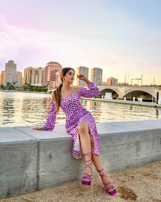 Roopsi Bansal in a purple leopard-print off-shoulder midi dress, seated on a seawall with the West Palm Beach skyline and a bridge glowing at sunset behind her