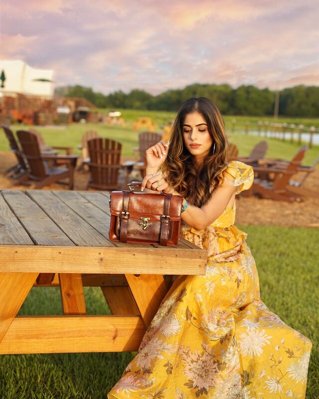 Roopsi Bansal in a yellow floral maxi dress, seated at a wooden picnic table with a vintage brown leather briefcase, Adirondack chairs and open grounds at Westgate River Ranch in the background