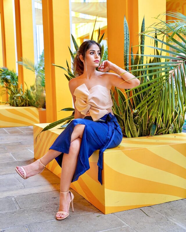 Roopsi Bansal in a bow-front nude top and royal blue wrap skirt, demonstrating relaxed hand poses in a bold yellow colonnade lined with snake plants