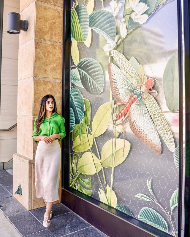 Roopsi Bansal in a green silk blouse and cream silk midi skirt from MOTF, leaning against a pillar beside a large botanical mural of a grasshopper on leaves