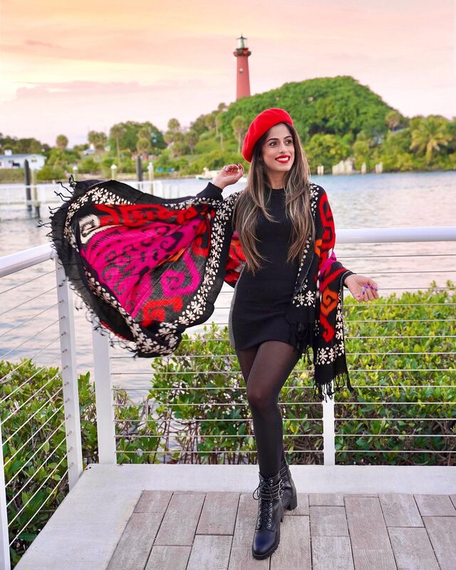 Roopsi Bansal in a red beret and black dress, holding out a colorful printed wool shawl, standing on a deck overlooking the Jupiter Inlet Lighthouse at sunset