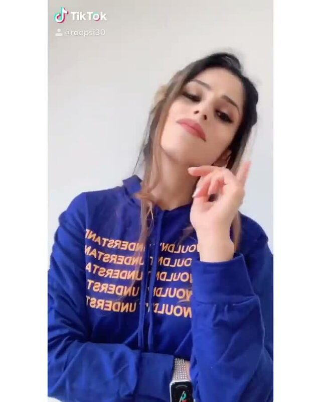 Roopsi Bansal in a blue hoodie printed with repeating text, filming a TikTok outfit-change video themed around countries
