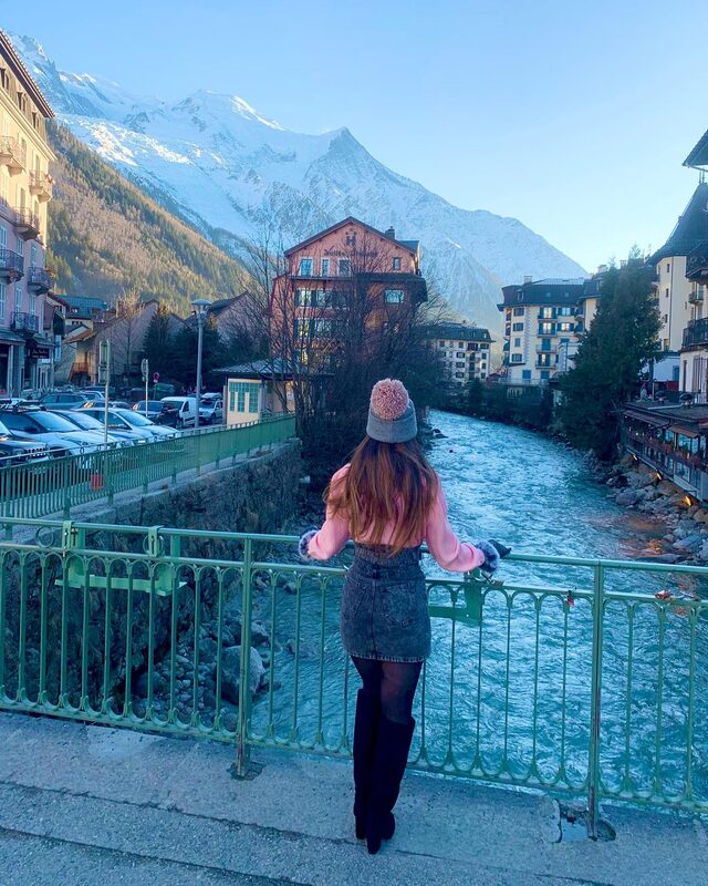 Standing on a green iron bridge over a glacial river in Chamonix, France, wearing a pink pom-pom beanie, pink sweater, denim skirt, and tall black boots, with the snow-capped Mont Blanc massif rising behind the town