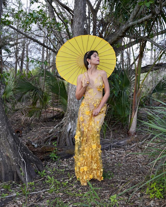 Roopsi Bansal in a yellow 3D floral-appliqué maxi dress from Dress the Population, holding a yellow parasol, standing among trees and palmetto in a Florida woodland