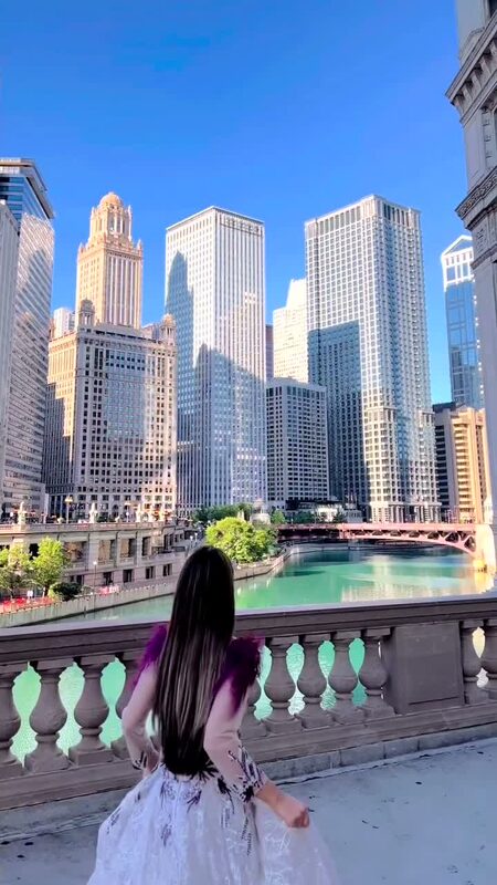 Roopsi Bansal from behind in a white lace gown with mauve feather sleeve trim, overlooking the Chicago Riverwalk skyline and teal-green river