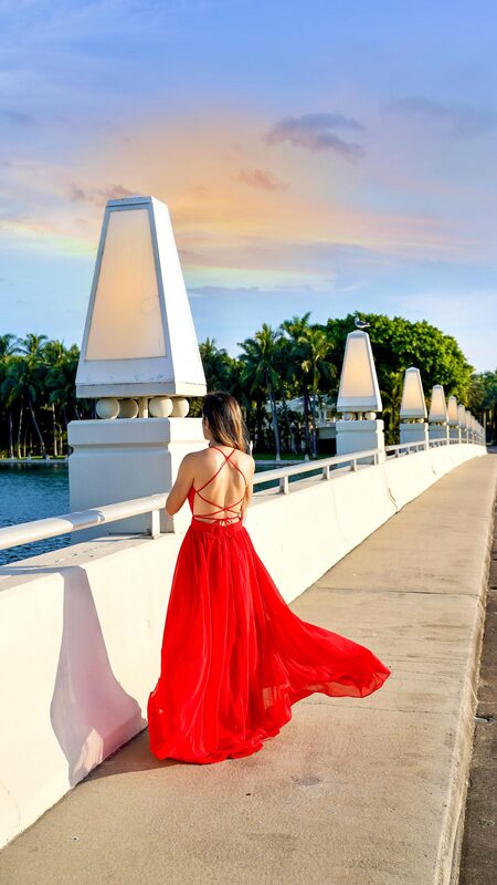 Roopsi Bansal in a long red backless gown standing on a waterside bridge at sunset, her back to the camera as she looks out toward the water