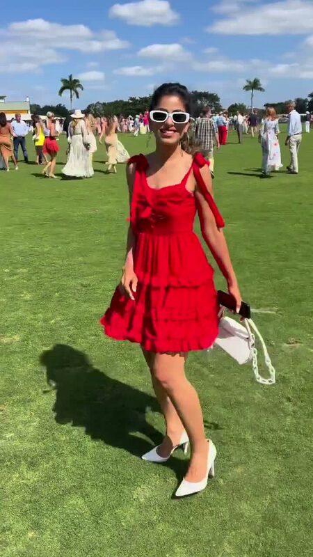 Roopsi Bansal in a red ruffled mini dress with bow straps from ROMWE and white sunglasses, standing on the polo grounds at the Winter Equestrian Festival with a crowd behind her