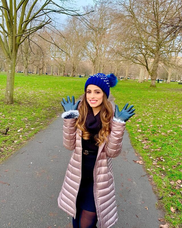 Standing on a park path in a blue rhinestone pom-pom beanie, blush puffer coat, navy scarf and dress with a Gucci belt, wearing blue leather gloves, surrounded by bare winter trees
