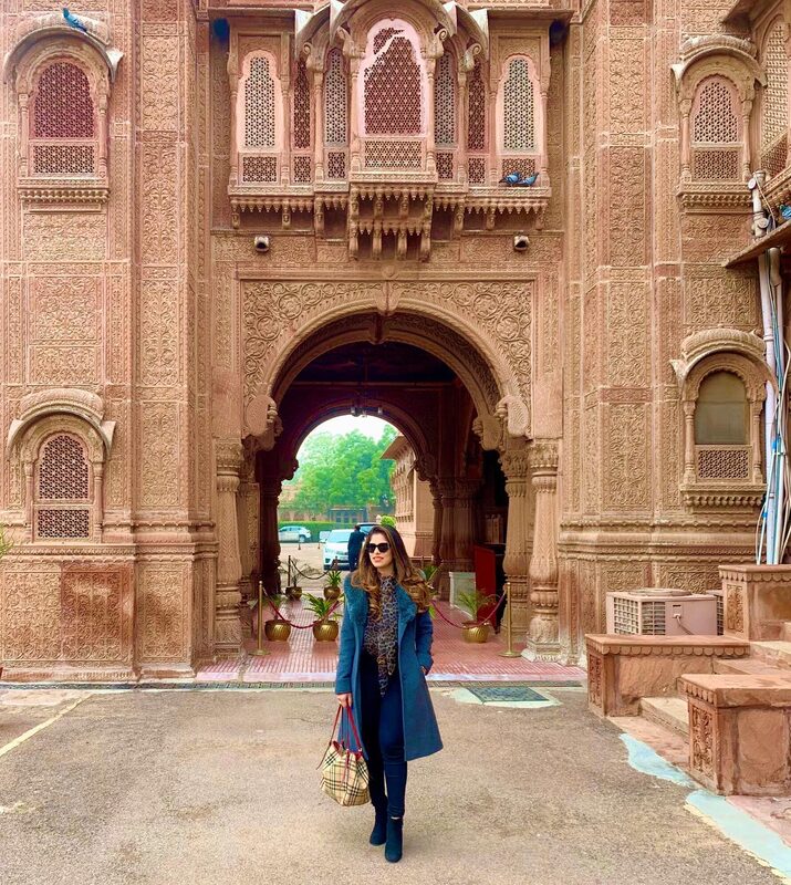 Standing before the ornately carved pink sandstone archway of Laxmi Niwas Palace in Rajasthan, India, wearing a teal coat, leopard-print scarf, and carrying a Burberry check tote
