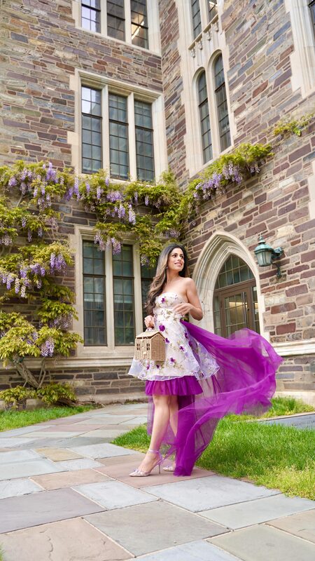 Roopsi Bansal in a floral dress with a dramatic purple tulle train, wisteria cascading down a stone wall behind her