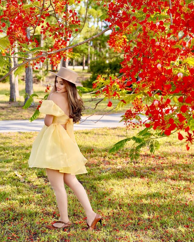 Roopsi Bansal in a yellow off-shoulder puff-sleeve organza mini dress and a beige Dahlia Boater hat from Gigi Pip, standing beneath a tree in full red bloom