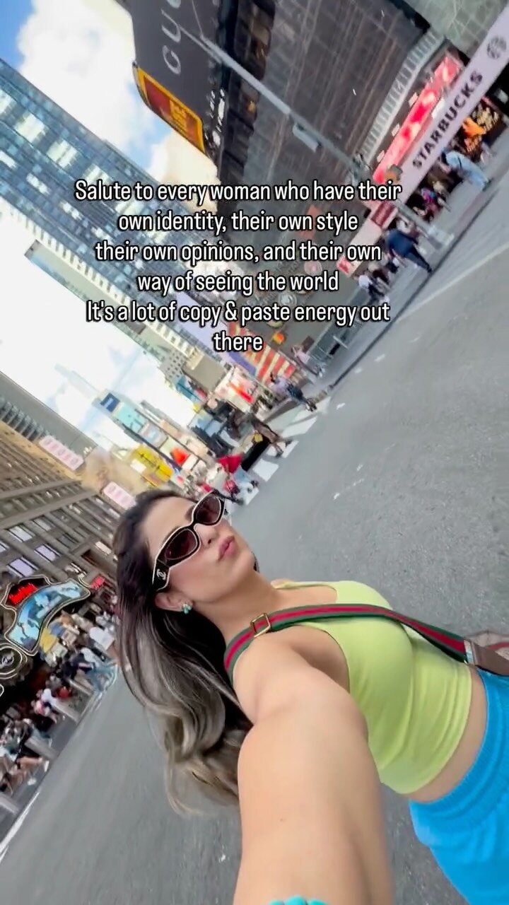 Roopsi Bansal taking a selfie in Times Square wearing a neon lime crop top, a striped crossbody strap, and sunglasses, with on-screen text saluting every woman with her own identity