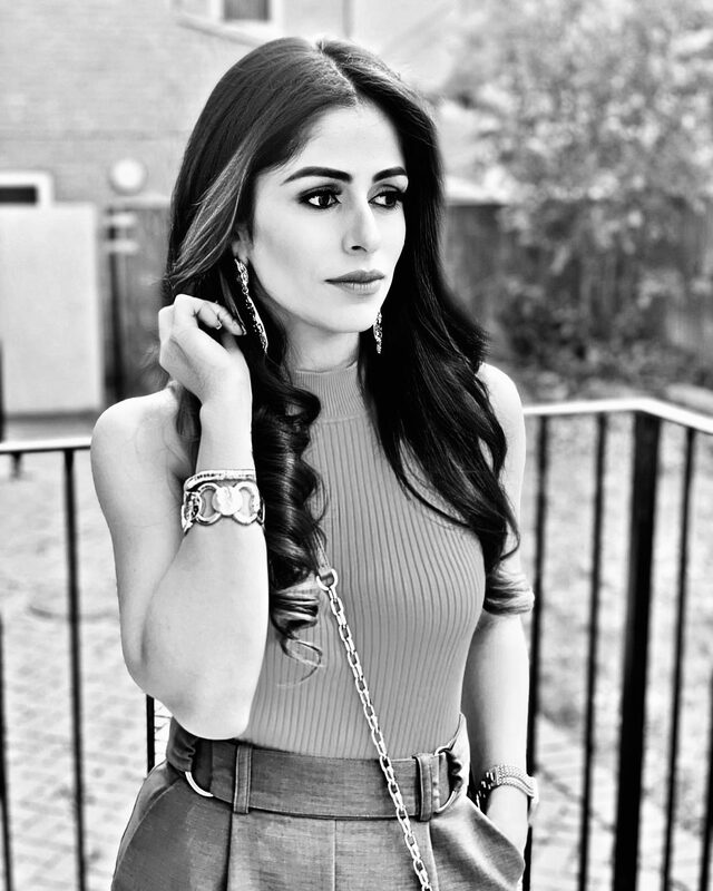 Black-and-white portrait wearing a ribbed sleeveless top and a pleated skirt with a Gucci logo belt, one hand raised to her hair, standing by a black metal railing