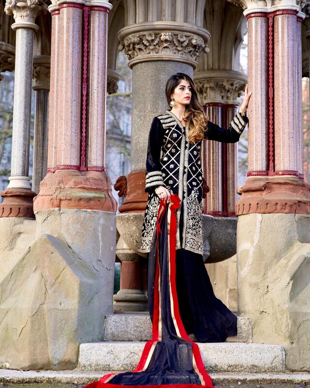 Roopsi Bansal in a black and gold embroidered long jacket with a red, black and cream striped train, posing on the steps of an ornate stone memorial