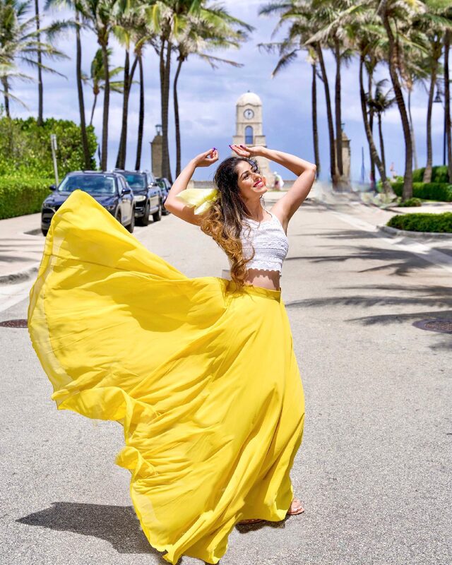Roopsi Bansal twirling in a bright yellow maxi skirt and white crochet top on Worth Avenue, palm trees framing the historic clock tower behind her in West Palm Beach