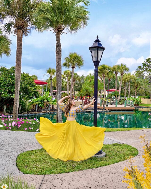 Roopsi Bansal in a plaid off-shoulder crop top and a swishing yellow maxi skirt, posing beside a lamppost next to a lagoon-style pool at Wyndham Orlando Resort