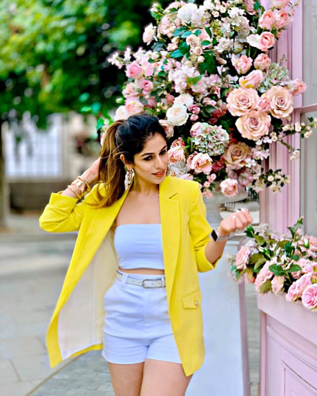 Standing in front of a wall of pink and white roses at Peggy Porschen's pink London bakery, wearing a yellow blazer over a pale blue tube top and white shorts