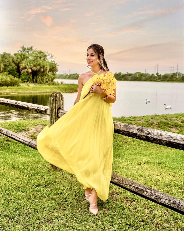 Roopsi Bansal in a flowing yellow halter dress, leaning on a wooden fence by a lake at sunset with two swans swimming in the background
