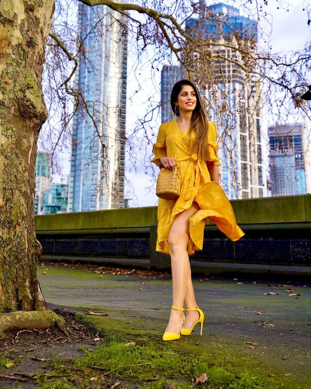 Roopsi Bansal in a ruffled yellow wrap dress and yellow ankle-strap heels, mid-stride in front of a row of glass skyscrapers