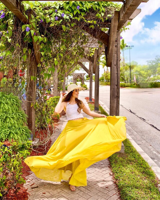 Roopsi Bansal in a white crochet top and flowing yellow maxi skirt with a straw hat, twirling under a flower-covered wooden pergola