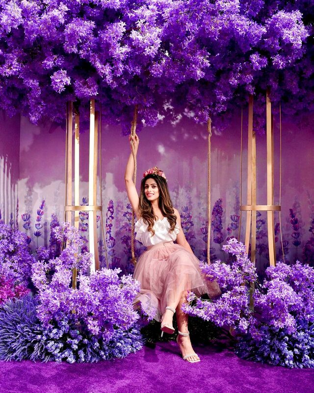 Roopsi Bansal in a white ruffle top and blush tulle midi skirt, seated on a garden swing beneath a canopy of hanging purple flowers at a floral installation